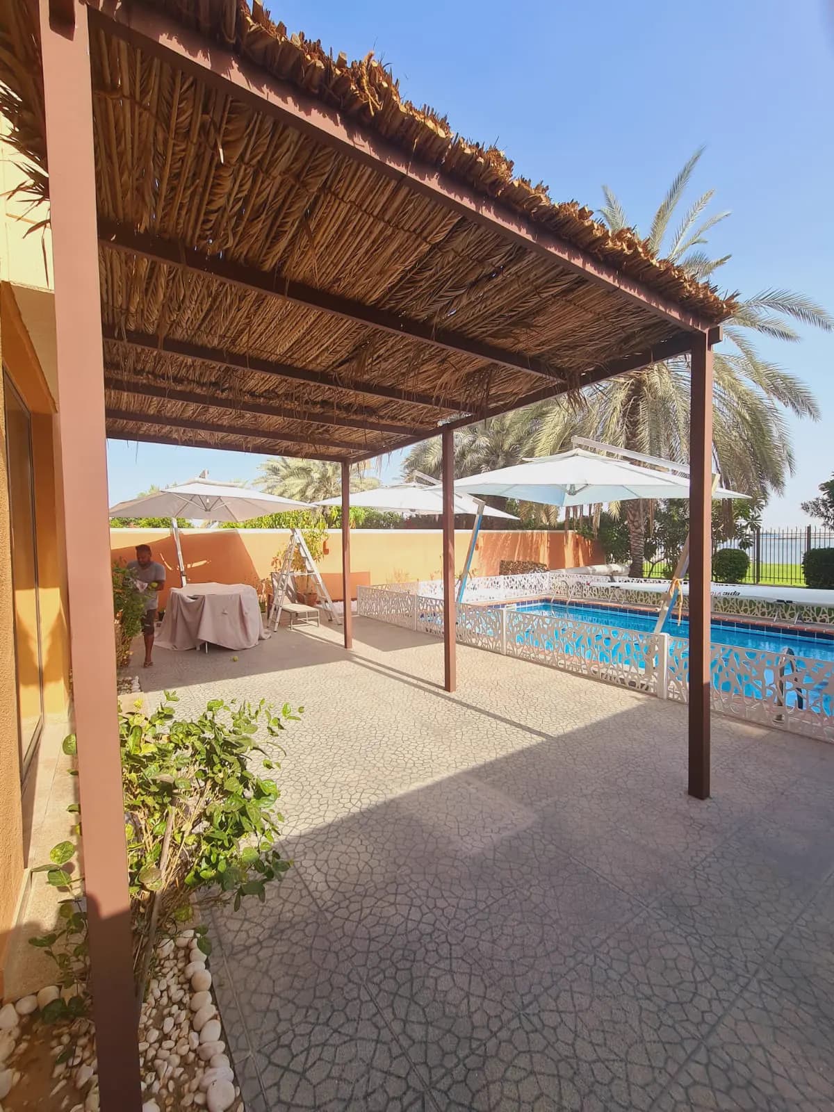 Timber pergola with palm-thatch roof shading a poolside terrace and sun loungers