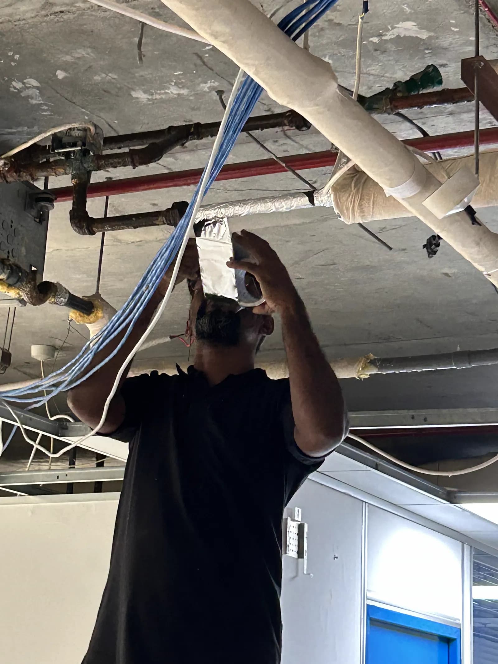 Technician terminating low-current cabling at a ceiling device beside ducting and pipework