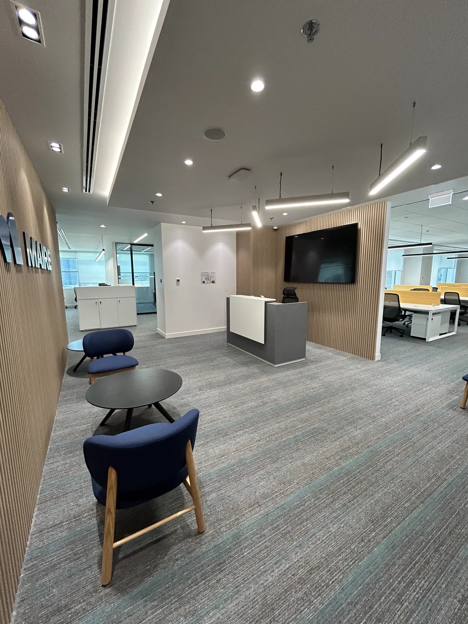 Office lounge with wall-mounted display, low seating and workstations beyond