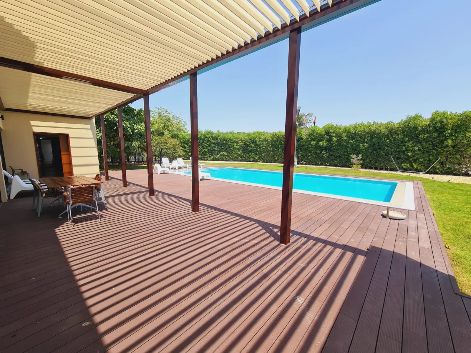 Shaded timber deck under a pergola looking onto a swimming pool and hedge